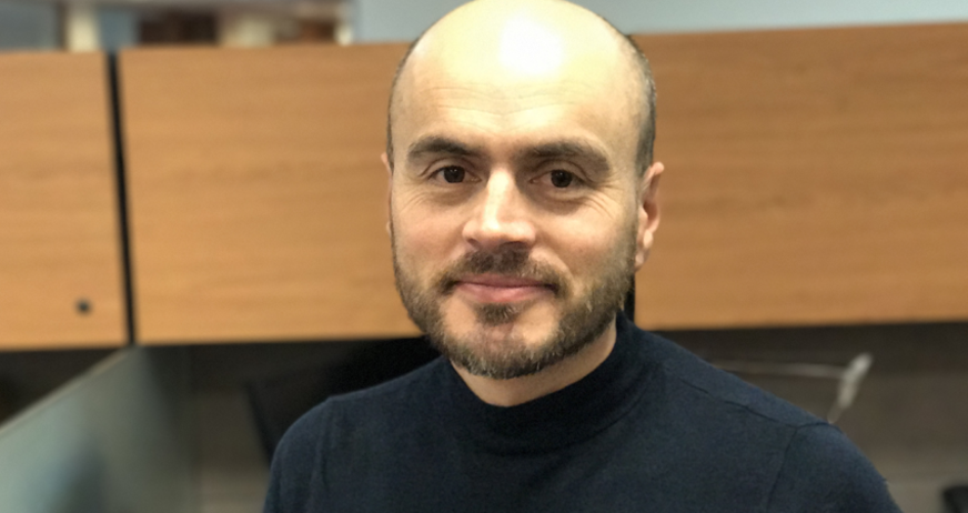 A man with a beard and a shaved head wearing a black sweater stands in front of wooden dividers.